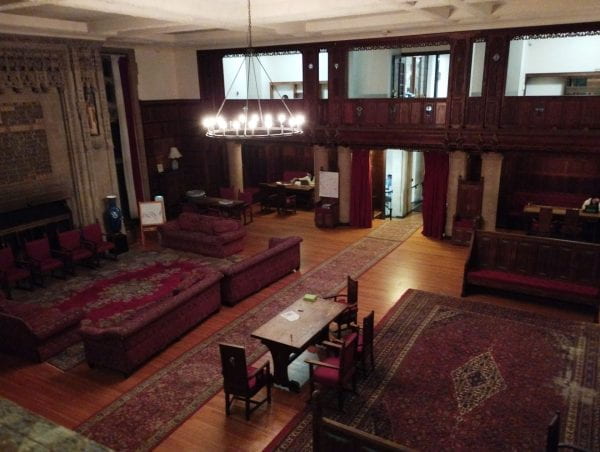 A view overlooking the great hall, which includes wood paneled walls, large couches, some tables, and large stately rugs