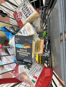 Image of a shopping cart basket full of different kinds of cheese like brie and provolone.