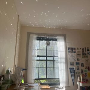 Image of a dorm room window with a disco ball hanging from the top sending shimmering light dancing across the walls.