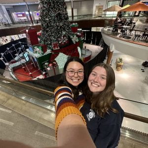 Jade and a friend standing in front of a Christmas tree in the center of a mall atrium.
