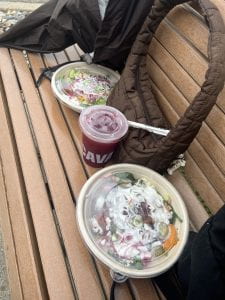 Image of takeout food and a drink from Cava on a park bench amongst purses and coats.