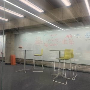 A concrete room with colorful yellow hightop chairs and tables against a backdrop of a wall covered in a whiteboard.
